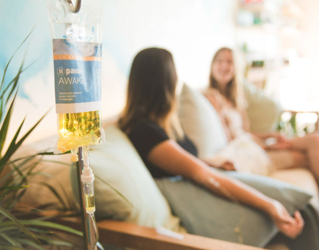 IV drip bag with two women relaxing in the background during IV therapy