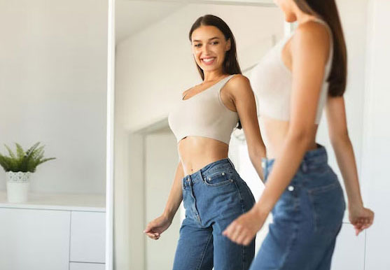 Woman smiling at her reflection while trying on jeans after weight loss